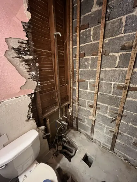 Shower area stripped to the wood framing and concrete block wall, with the old plumbing exposed and a hole in the floor for the drain.